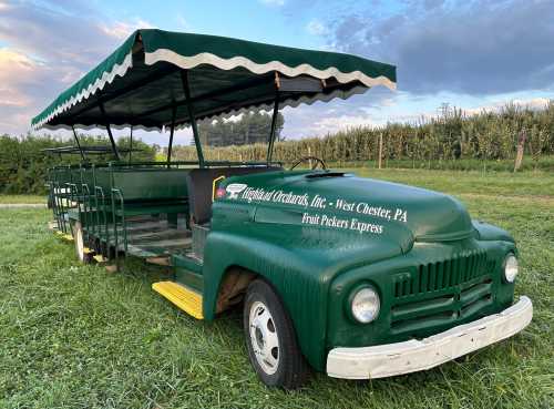 A green fruit-picking tram with a canopy, parked in an orchard with trees in the background under a cloudy sky.