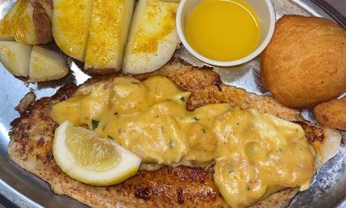 A plate featuring grilled fish topped with creamy sauce, lemon slices, baked potatoes, a roll, and a cup of butter.