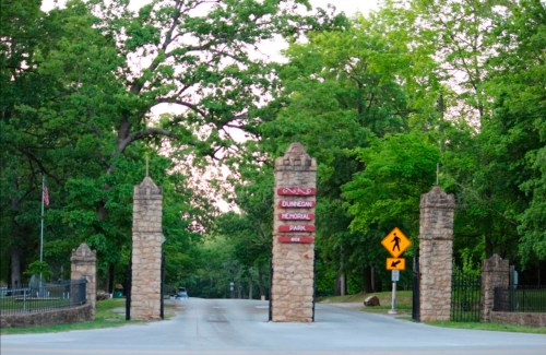 Stone entrance gates to Dunedin Memorial Park, surrounded by lush green trees and a clear sky.