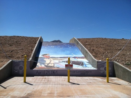 A mural depicting a historical scene on a sloped concrete structure, with mountains in the background and a clear blue sky.