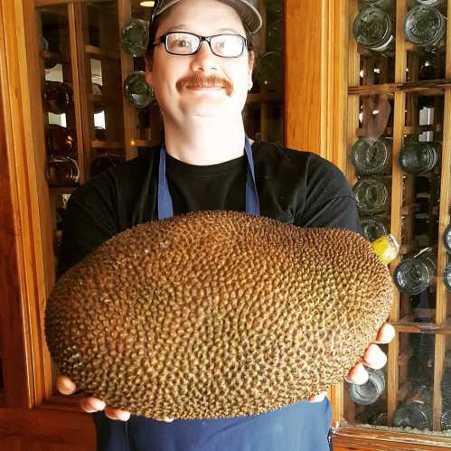 A person with glasses and a cap holds a large, spiky jackfruit, smiling in a wooden-paneled setting.