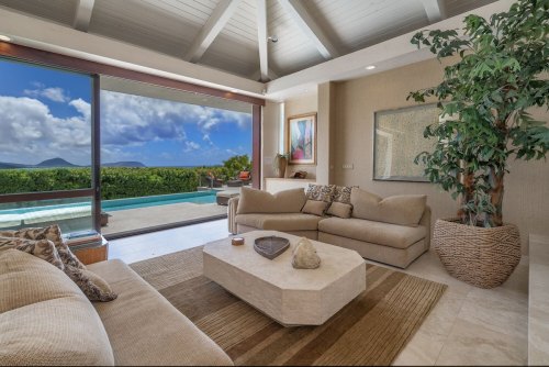A modern living room with beige sofas, a coffee table, and a view of a pool and landscape through large windows.