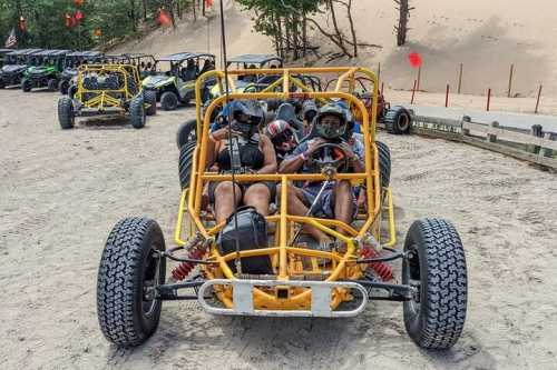 Two people in a yellow dune buggy, wearing helmets, ready to drive on a sandy track with other buggies in the background.