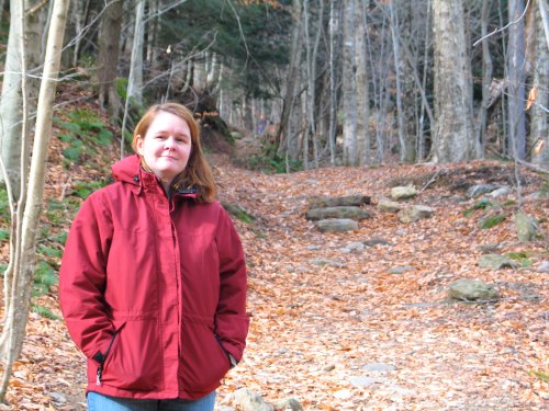 A woman in a red jacket stands on a leaf-covered forest path, surrounded by trees in autumn.