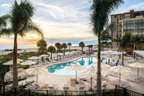 A serene pool area with lounge chairs and umbrellas, surrounded by palm trees, overlooking a beach at sunset.