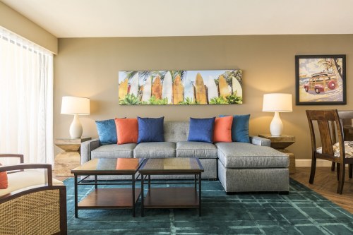 Cozy living room with a gray sofa, colorful pillows, coffee tables, and tropical artwork on the wall.