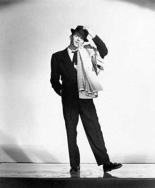 A stylish man in a suit and hat poses confidently, holding a scarf, against a simple backdrop.