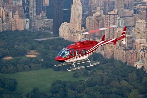 A red and white helicopter flying over a city skyline and a green park.