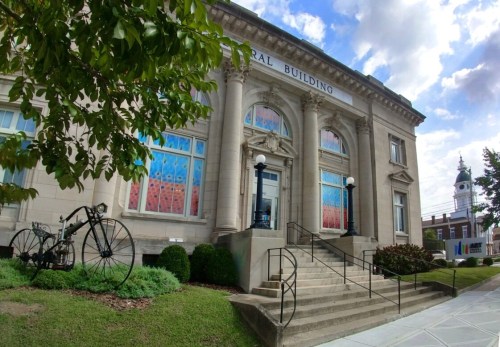 Historic building with large windows, decorative facade, and a vintage bicycle in front, surrounded by greenery.
