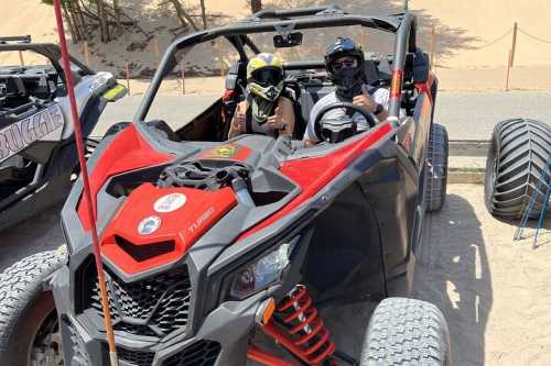 Two people wearing helmets and masks sit in a red off-road vehicle on sandy terrain, ready for an adventure.