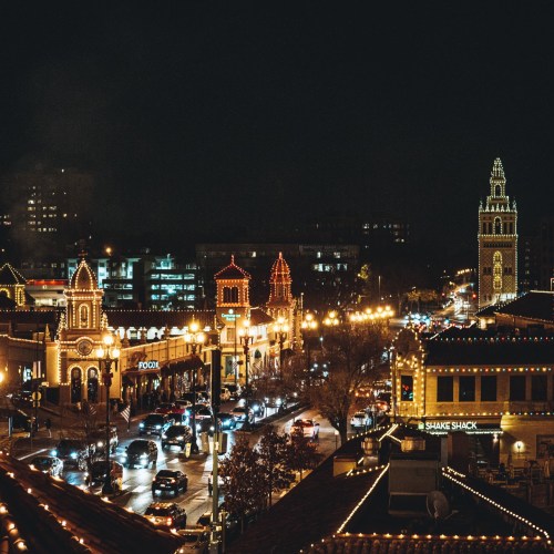 A vibrant city street at night, illuminated by festive lights and bustling with cars and pedestrians.