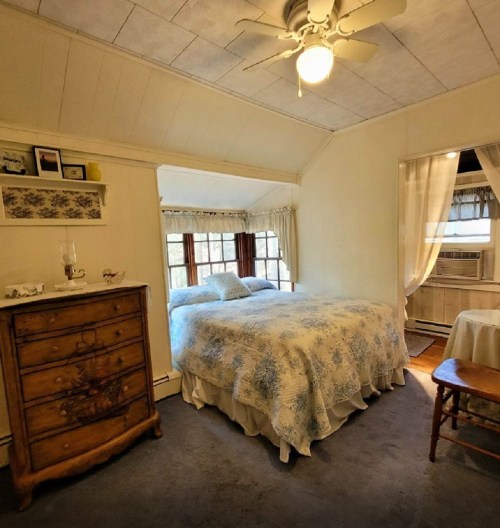 Cozy bedroom with a floral bedspread, wooden dresser, and natural light from windows. Soft, inviting decor.