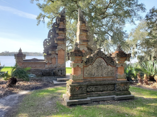 Two ornate stone structures with intricate carvings, surrounded by greenery and overlooking a body of water.
