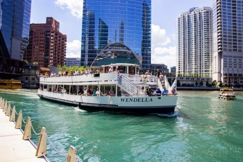 A tour boat named "Wendella" navigates a river surrounded by tall buildings on a sunny day.