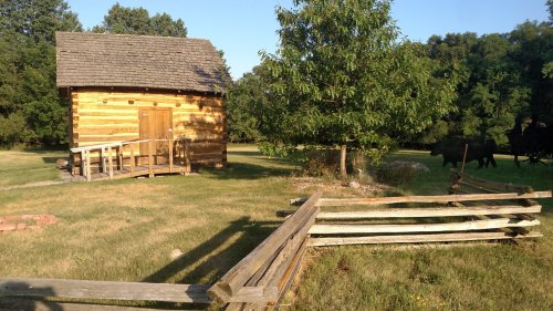 A rustic wooden cabin surrounded by green grass and trees, with a wooden fence in the foreground.