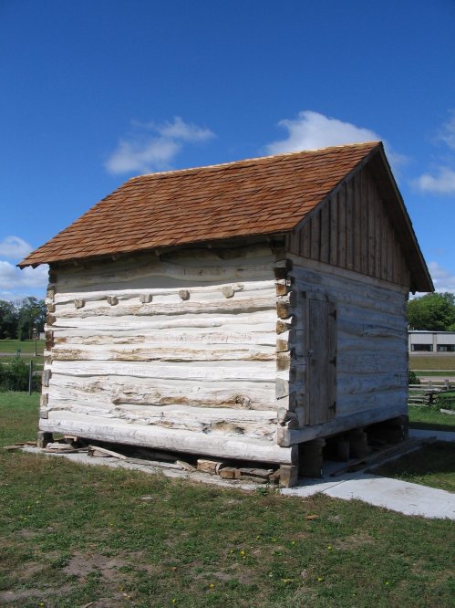 A small wooden cabin with a sloped roof, set against a blue sky and grassy landscape.