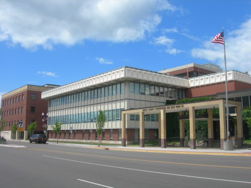Modern brick and glass building with an American flag, set against a blue sky with clouds. Street view.