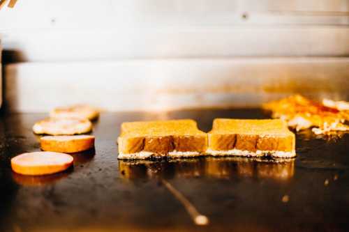 Two slices of bread grilling on a flat surface, with some circular meat slices nearby.