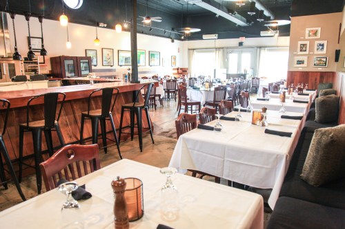 Interior of a restaurant featuring wooden tables, chairs, a bar area, and large windows with natural light.