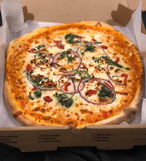A freshly baked pizza with cheese, red onions, spinach, and diced tomatoes in a takeout box.