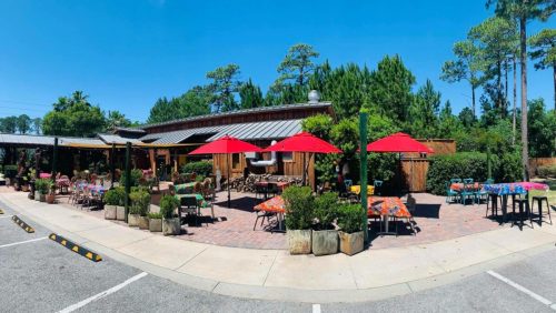 Outdoor dining area of a restaurant with colorful tables, red umbrellas, and lush greenery under a clear blue sky.