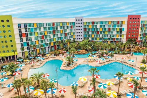 Colorful hotel exterior with a large pool area, surrounded by palm trees and umbrellas, under a bright blue sky.