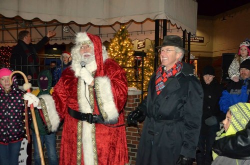 Santa Claus speaks into a microphone beside a man in a hat, with a crowd of children and festive lights in the background.