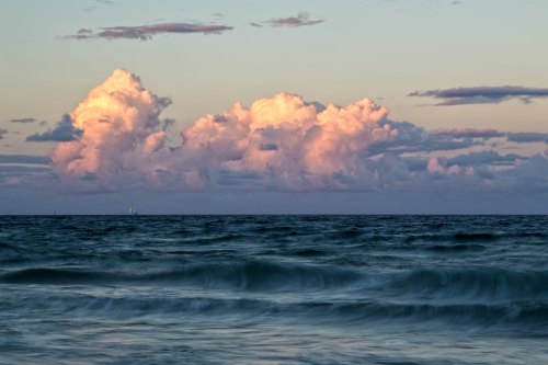 A serene seascape at sunset, featuring soft pink clouds above gentle ocean waves.
