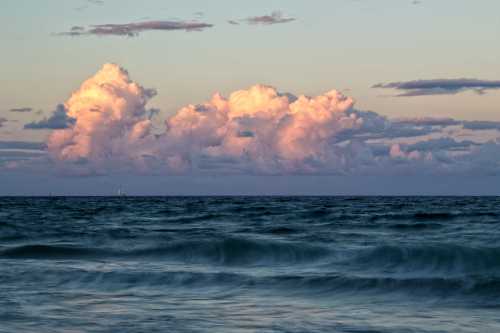 A serene seascape at sunset, featuring soft pink clouds above gentle ocean waves.