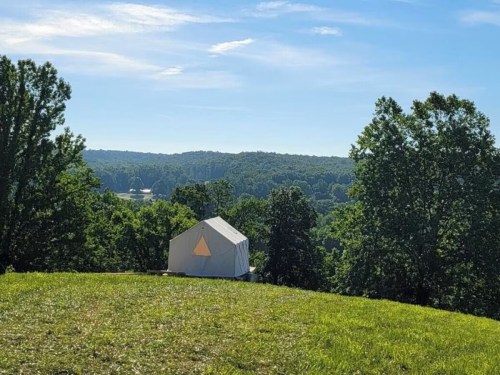 A white tent sits on a grassy hill, surrounded by lush green trees and a scenic view of rolling hills in the background.