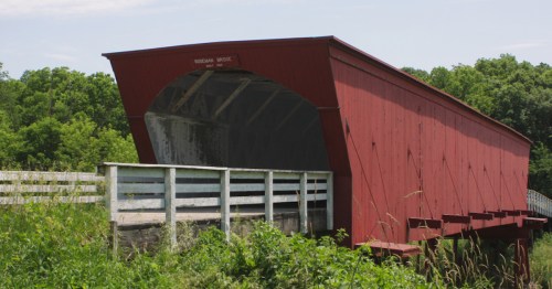 A red covered bridge spans a small stream, surrounded by lush greenery and a white wooden railing.