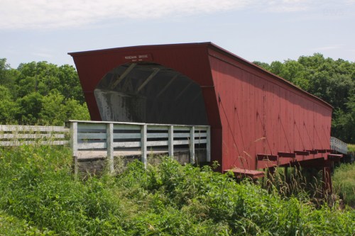A red covered bridge stands over a grassy area, surrounded by trees and a white fence under a clear sky.