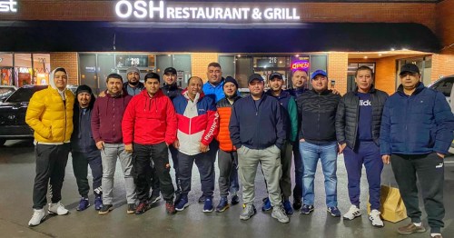 A group of men posing together outside OSH Restaurant & Grill, smiling and wearing casual winter clothing.