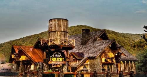 A rustic lodge with a water tower, featuring a sign for "Breakfast Camp," set against a mountainous backdrop.