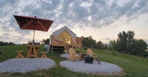 A cozy glamping tent on a grassy hill, with a picnic table, chairs, and an umbrella, under a cloudy sky at sunset.