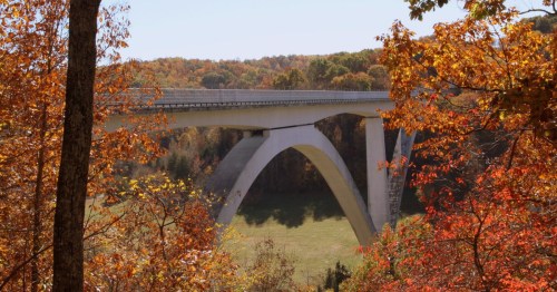 A modern concrete bridge arches over a valley, surrounded by vibrant autumn foliage.