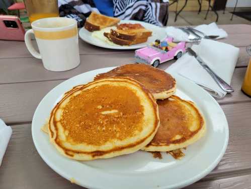 Three golden pancakes on a white plate, with toast and a pink toy car in the background on a wooden table.