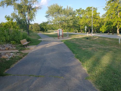 A paved path in a park with benches, trees, and a signpost, leading towards a road in the distance.