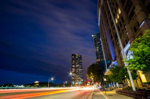 City skyline at night with light trails from traffic, featuring tall buildings and a cloudy sky.