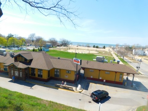 A view of a beachside building with a parking lot, grassy area, and a glimpse of the lake in the background.