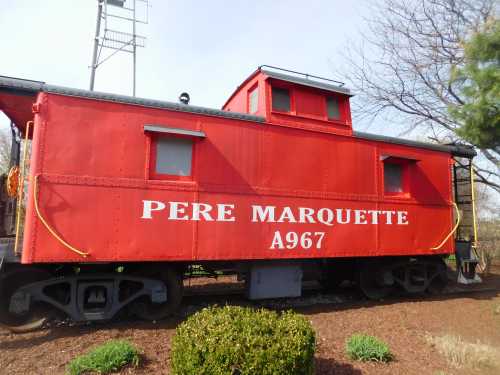 A bright red caboose labeled "Pere Marquette A967" parked among greenery and trees.