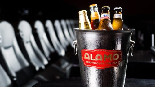 A metal bucket filled with various beer bottles, featuring the Alamo Drafthouse Cinema logo, set against empty theater seats.