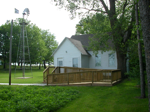 A small white house with a wooden deck, surrounded by green grass and trees, with a windmill in the background.