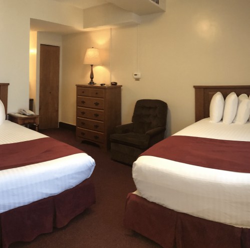 A cozy hotel room with two beds, a dresser, a chair, and warm lighting.