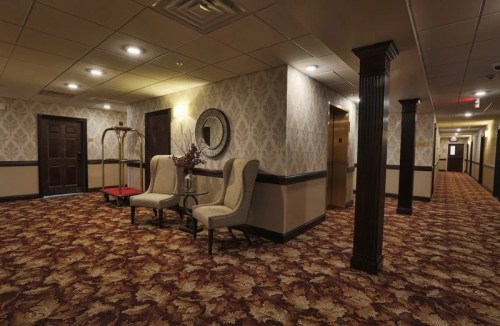 A hotel hallway featuring patterned carpet, two chairs, a mirror, and an elevator in the background.