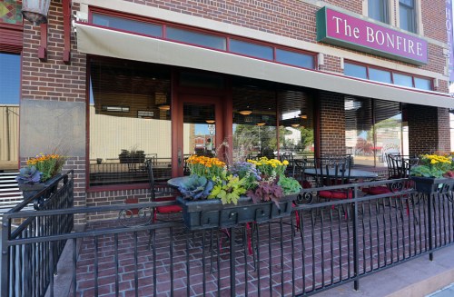 Exterior view of The Bonfire restaurant, featuring a patio with flowers and empty tables under a canopy.