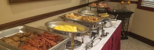A buffet table featuring scrambled eggs, bacon, sausage, potatoes, and pastries in chafing dishes.