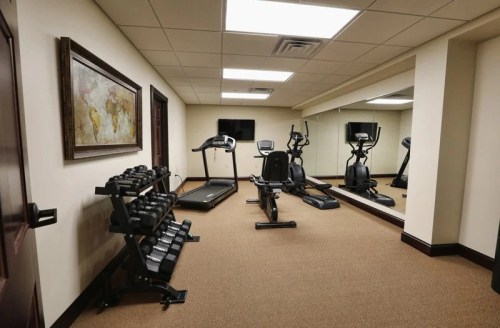 A fitness room featuring a treadmill, elliptical, weights, and a large mirror, with soft lighting and carpeted floor.