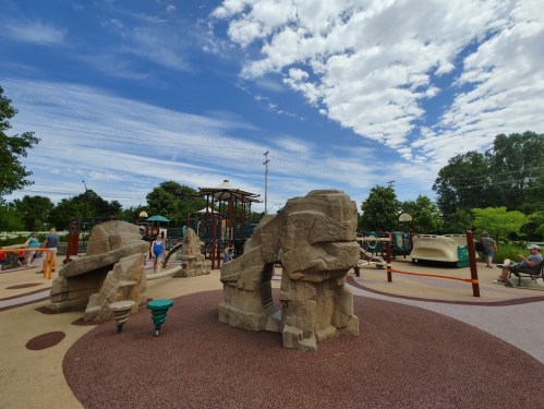 A playground featuring rock formations, climbing structures, and colorful equipment under a blue sky with scattered clouds.