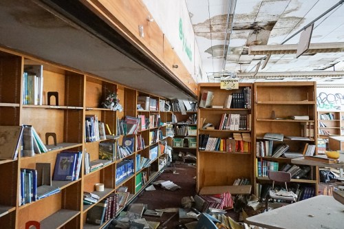Abandoned library with disorganized bookshelves, scattered debris, and a deteriorating ceiling.